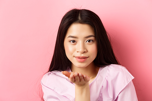 Beautiful asian girl with natural makeup, blowing air kiss at camera with sensual gaze, standing romantic against pink background.