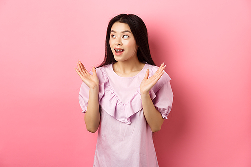 Excited asian girl clap hands and looking left, watching performance and applause, standing in dress on pink background.