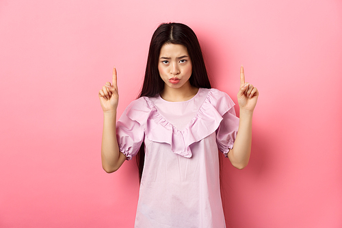 Sad and moody asian girl look from under forehead and sulking, pointing fingers up and complaining, staring jealous or upset, standing against pink background.