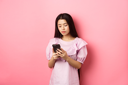 Teenage asian girl chatting on mobile phone, look at smartphone screen with serious face, standing against pink background.