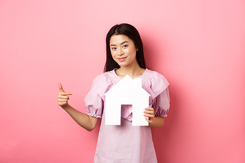 Real estate and insurance concept. Beautiful asian woman pointing at paper house cutout, showing agency logo, standing in dress on pink background.