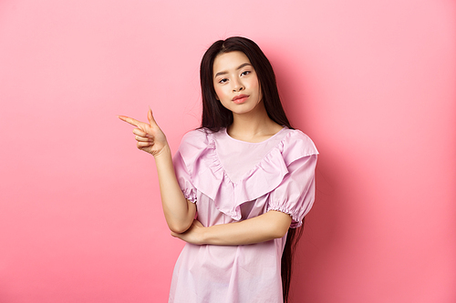 Young smug asian girl looking cool and pointing finger left at logo, advertising product on pink romantic background.
