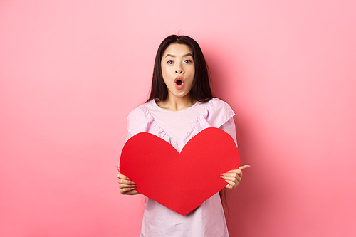 Valentines day concept. Surprised asian teen girl say wow, showing big red heart card, receive gift from secret admirer or lover, standing in romantic dress on pink background.