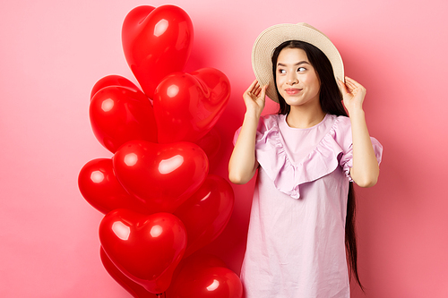 Tender and romantic asian teen girl wearing straw hat and dress on date, standing near valentines day heart balloons and looking aside with dreamy smile, pink background.