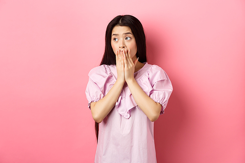 Shocked and worried asian woman looking aside at empty space with concerned face, covering mouth with hands, standing in dress on pink background.