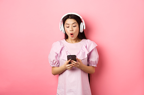 Excited asian girl looking at smartphone screen amused, say wow, listening music in wireless headphones, standing against pink background.