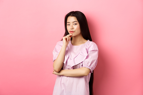 Hmm, interesting. Pensive and cunning asian girl having idea, plan to do something,  and touching chin with thoughtful face, standing on pink background.