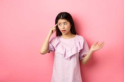 Are you stupid. Shocked asian woman pointing at head and complaining, scolding someone crazy or strange, look aside, standing against pink background.