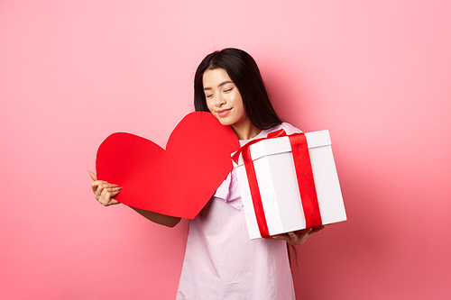 Valentines day concept. Romantic asian teen girl receive gifts on white day from lover, hugging big red heart card and present in box, smiling sensual, pink background.