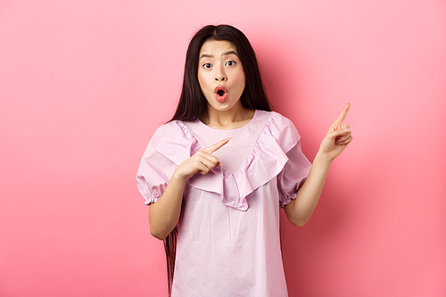 Excited asian woman look amazed, pointing right and gasping fascinated, checking out advertisement, standing on pink background.