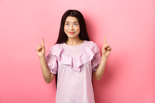 Beautiful smiling asian teen girl pointing fingers up, looking at advertisement with happy face, standing on pink background.