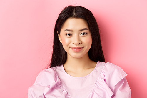 Close-up of beautiful asian woman looking happy, smiling cute at camera, standing in dress against pink background.