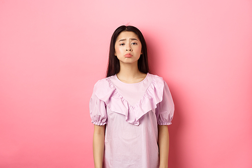 Sad whining asian girl pouting and frowning, looking upset about something unfair, complaining, standing in dress against pink background.