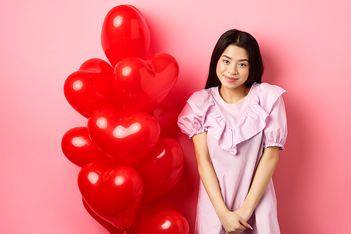 Cute asian girl in dress looking shy and smiling, standing modest near valentines day balloons, blushing on romantic date, , pink background.