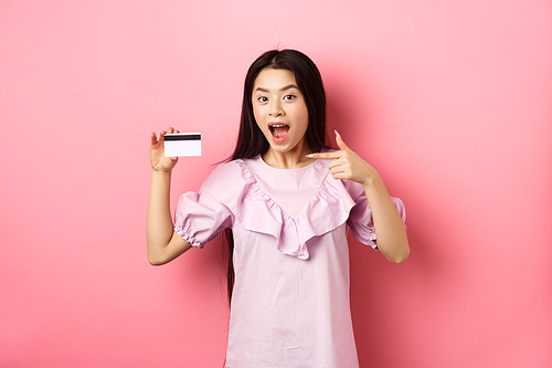 Happy cute asian woman showing plastic credit card, recommending bank special deal, standing against pink background.