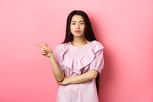 Skeptical chinese girl pointing finger left, looking unamused and serious, showing offer, standing in dress on pink background.