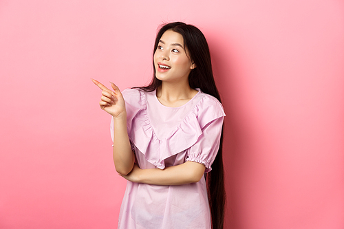 Excited smiling asian woman in dress, pointing and looking left at logo, checking out promotion, standing on pink background.