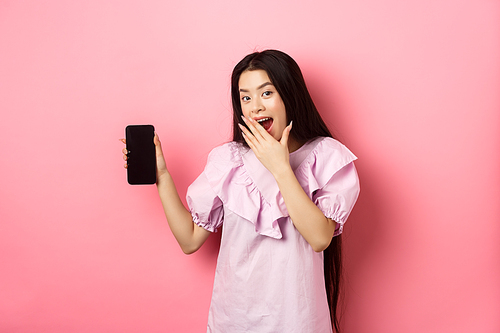 Cheerful asian teen girl showing empty phone screen, laughing and covering mouth with hand, standing in dress against pink background.