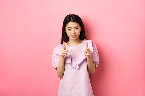 Sassy asian woman pointing at camera, smiling and inviting you, beckon or praise person, standing on pink background.