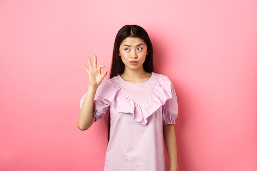 Sassy asian woman in dress show okay sign, looking aside, assure everything good, guarantee all OK, standing pleased on pink background.
