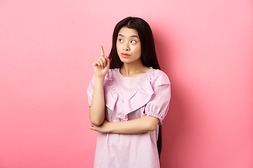 Beautiful asian girl raising finger and looking left at logo, having an idea, found solution, standing against pink background.