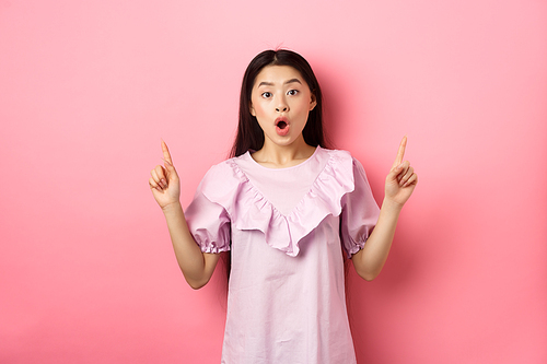 Excited asian teen girl pointing fingers up, saying wow and showing promo deal, standing in dress against pink background.