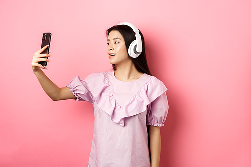 beautiful asian girl Vlog taking selfie in wireless headphones, make photo for social media on smartphone, standing in dress against pink background.