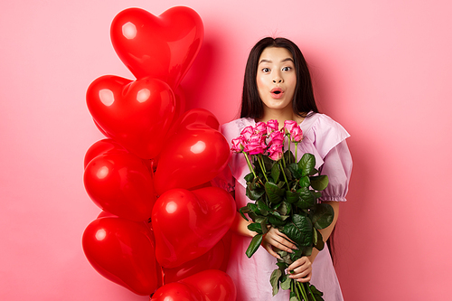 Surprised asian girl in dress standing near valentines day heart balloons and say wow at camera, holding flowers bouquet from lover, romantic date with roses, pink background.