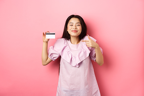 Cheerful and cute asian woman smiling satisfied, showing plastic credit card, standing in dress against pink background.
