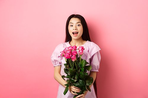 Korean teen girl in dress having romantic date on Valentines day, holding bouquet of roses and looking surprised at camera, receive gift on date from lover, pink background.