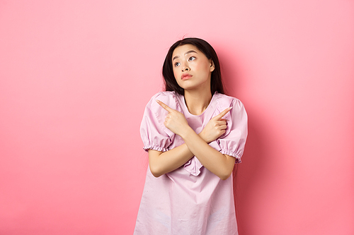Indecisive asian woman cant decide, pointing fingers sideways and look pensive aside, making choice, standing confused on pink background.