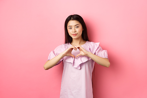 Valentines day concept. Beautiul japanese teen girl show heart gesture and smiling cute, express love and sympathy to lover, standing in romantic dress on pink background.