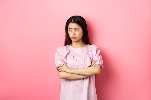 Sad and gloomy asian woman look away at logo, sulking from unfair situation, cross arms on chest disappointed, standing against pink background.