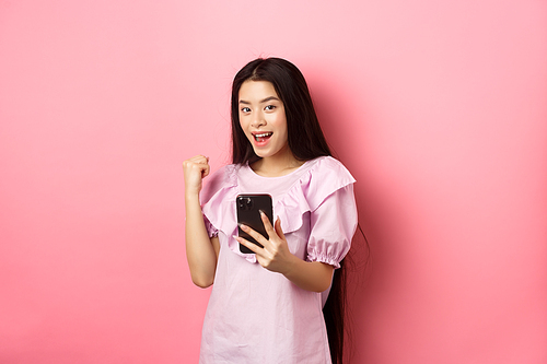 Online shopping. Satisfied asian girl winning on mobile phone, say yes and make fist pump, holding smartphone, standing against pink background.
