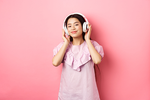 Beautiful asian woman listening music on wireless headphones, smiling pleased at camera, standing in dress against pink background.