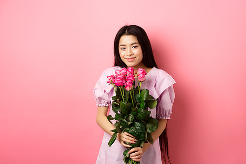 Romantic tender asian girl holding bouquet of roses, smiling cute at camera, having valentines date with lover, wearing dress, pink background.