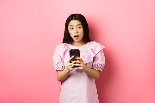 Online shopping. Surprised asian woman in cute dress, open mouth amazed after reading news on smartphone, standing on pink background.