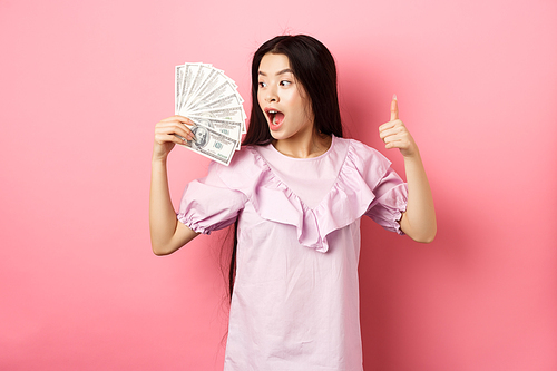 Excited asian girl looking at big money and make thumbs up, standing amazed with dollar bills against pink background.