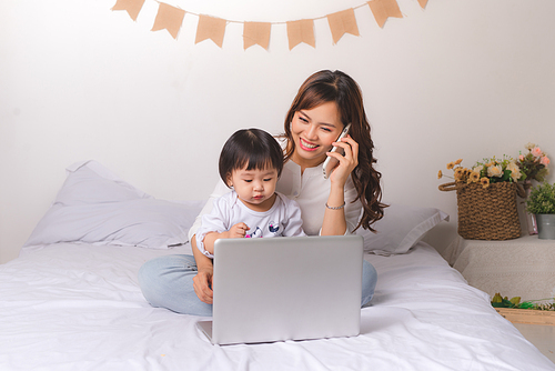 Asian lady in classic suit is talking on the mobile phone and working on laptop at home with her baby girl.