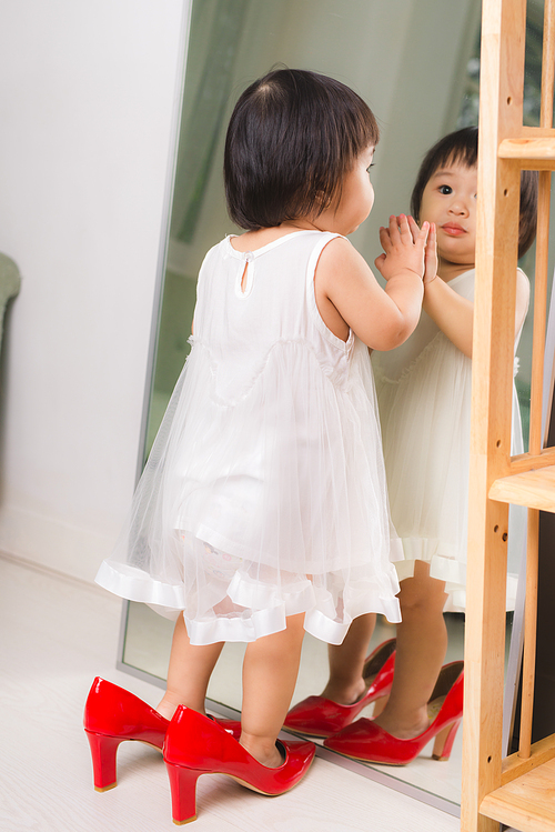 Cute funny little baby girl walks at home trying mom's red high heel shoes