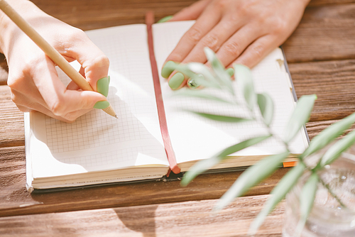 Unrecognizable young woman writing in notepad with a pen on wooden table, view of hands. Leaf plant in foreground.