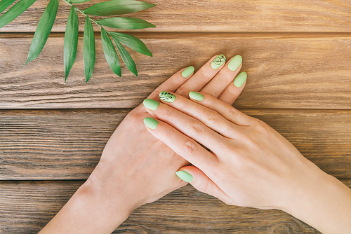 Female hands with art nails design and summer green stylish manicure near palm leaf on wooden background. Point of view in first person.