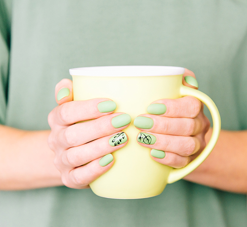 Close-up of female hands with art leaf nails design and trendy summer stylish green manicure holding a cup.