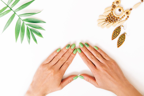 Female hands with art nails design and summer green color manicure near palm leaf and wooden handmade jewelry on white background. Point of view in first person.