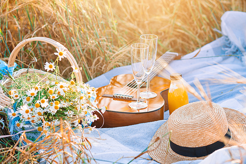 Closeup of picnic on nature in wheat field. Guitar, straw hat, wine with glasses and a basket of daisies