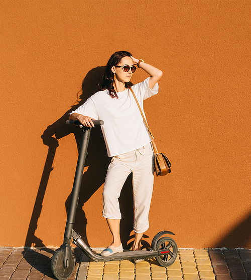 Beautiful young woman standing with electric scooter near the wall in street outdoor.