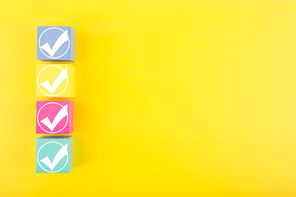 Four white checkmarks on multicolored toy cubes on bright yellow background with copy space. Concept of questionary, kids related checklist, to do list, planning, business or verification.