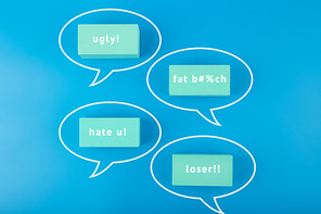 Creative cyberbullying concept. Minimal flat lay with negative, insulting messages in speech bubbles on blue background.