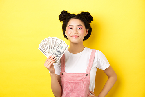 Shopping. Smiling asian girl with stylish makeup, showing dollar bills with pleased face, making money, standing on yellow background.