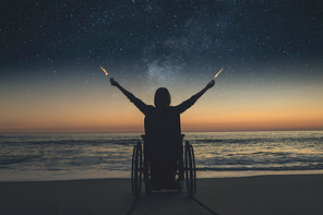 . woman in a wheelchair holding fire sparklers, at night on the beach